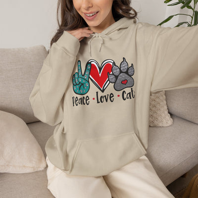 A woman wearing a beige hoodie with a graphic that reads 'Peace Love Cat' featuring a peace sign, red heart, and gray cat paw prints, sitting on a light gray couch.