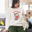 A woman sitting on a couch in a bright living room, wearing a white sweatshirt with a cartoon cat holding a red heart and the words 'ADOPT LOVE' in red. She is also wearing dark green pants and has a white and gray cat sitting on her lap.