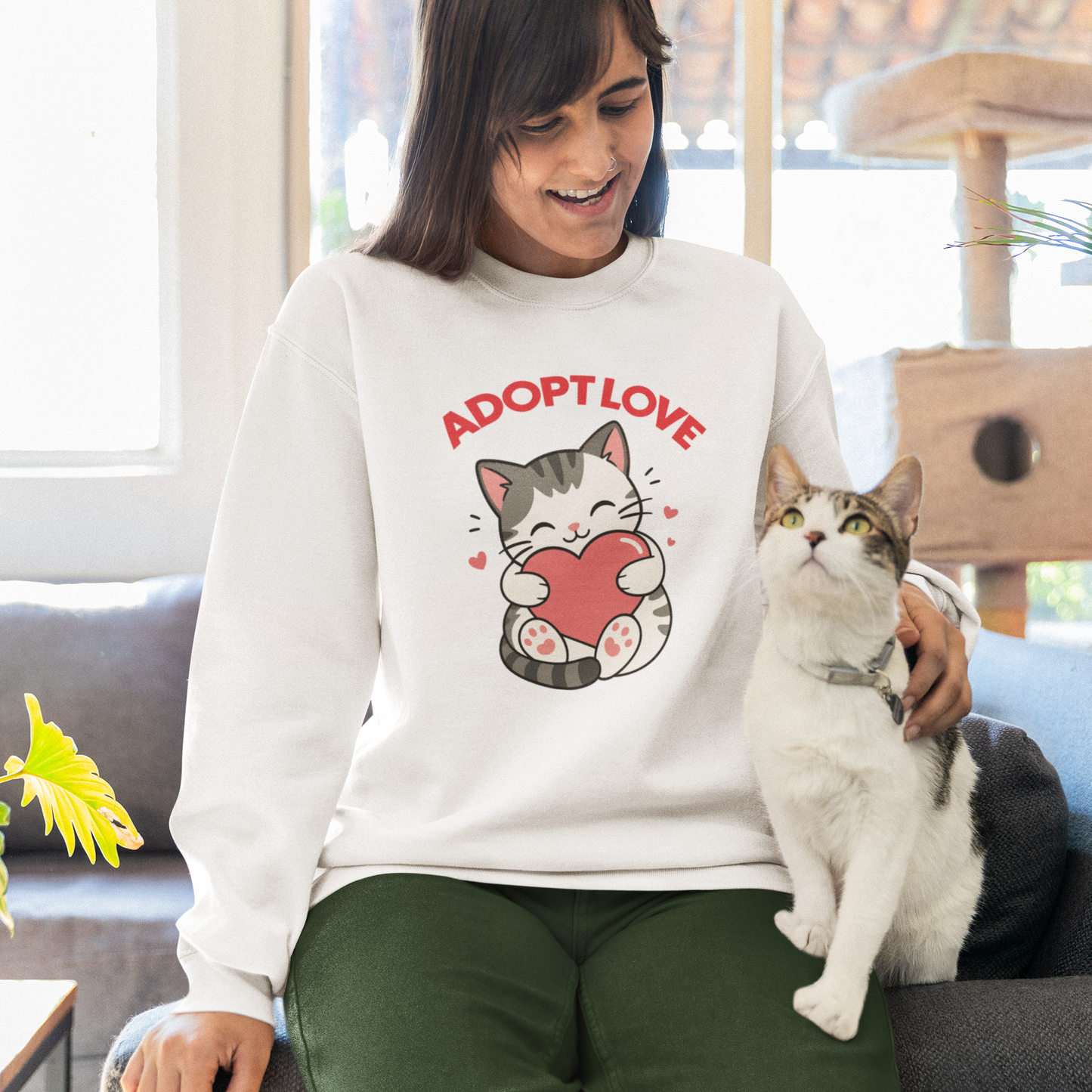 A woman sitting on a couch in a bright living room, wearing a white sweatshirt with a cartoon cat holding a red heart and the words 'ADOPT LOVE' in red. She is also wearing dark green pants and has a white and gray cat sitting on her lap.